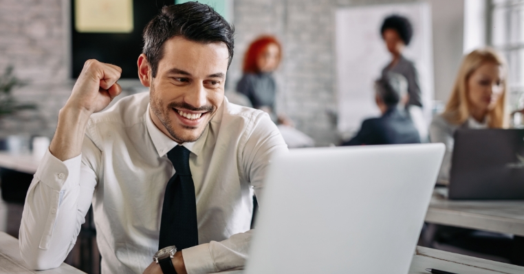 young-happy-businessman-using-computer-office-celebrating-good-news-there-are-people-background